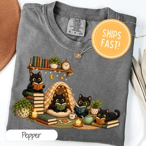 Könnte beinhalten: Ein anthrazitfarbenes T-Shirt mit einer Grafik von vier schwarzen Katzen, die Bücher lesen. Das Design zeigt ein Regal mit Büchern und einer Uhr, ein Zelt, Topfpflanzen, eine Teekanne und Kerzen. Das Shirt hat ein Comfort Colors-Etikett.