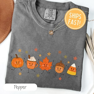 Könnte beinhalten: Ein anthrazitfarbenes Kurzarm-T-Shirt mit einem Grafikdruck von fünf lächelnden herbstlichen Charakteren: einem Kürbis, einer Tasse Kakao, einem Ahornblatt, einer Eichel und einem Zuckermais, umgeben von kleinen orangefarbenen und gelben Sternen.