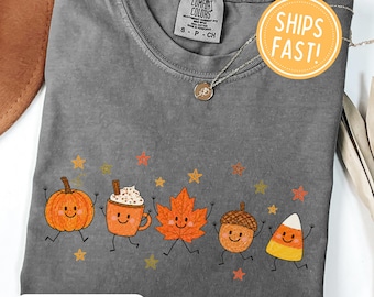Puede incluir: Una camiseta de manga corta gris carbón con un estampado gráfico de cinco personajes otoñales sonrientes: una calabaza, una taza de cacao, una hoja de arce, una bellota y un caramelo de maíz, rodeados de pequeñas estrellas naranjas y amarillas.