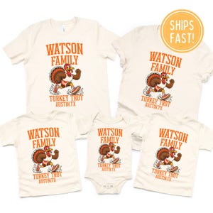 K&ouml;nnte beinhalten: Eine Kollektion aus cremefarbenen T-Shirts und einem Baby-Body mit einer orangefarbenen Grafik eines laufenden Truthahns. Der Text lautet WATSON FAMILY TURKEY TROT AUSTIN.TX. Das Design zeigt einen Truthahn mit Turnschuhen.