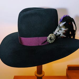 May include: A black fedora hat with a wide brim and a deep purple band. The hat is decorated with a decorative pin and a cluster of black, white, and purple feathers. The hat is displayed on a wooden stand.