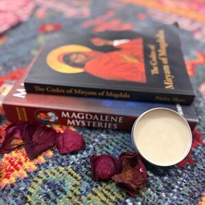 Peut inclure: Deux livres, "The Codex of Miryam of Magdala" et "Magdalene Mysteries", sont empilés avec un petit pot de crème argenté ouvert et des pétales de rose séchés. Les livres reposent sur une surface à motifs colorés.