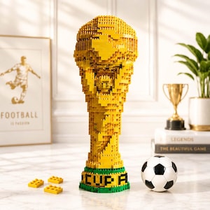 Könnte beinhalten: Eine goldene Trophäe aus ineinandergreifenden Kunststoff-Bausteinen mit einem texturierten, pixeligen Design. Der Sockel ist grün und trägt den schwarzen Schriftzug "CUP A". Rechts neben der Trophäe liegt ein kleiner schwarz-weißer Fußball.