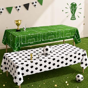 Könnte beinhalten: Zwei rechteckige Tischdecken für eine Party. Die obere zeigt ein grünes Fußballfeldmuster mit weißen Linien. Die untere hat ein schwarz-weißes Fußballmuster. Ein goldener Pokal, ein Becher und ein Fußball sind auf den Tischen platziert.