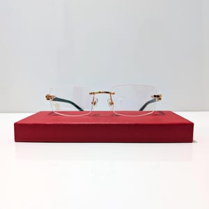 Könnte beinhalten: Randlose Brille mit goldfarbenen Akzenten und klaren Gläsern liegt auf einer roten rechteckigen Schachtel. Die Brille hat eine rechteckige Form und einen dünnen Goldrahmen. Die Schachtel hat eine einfarbige, tiefrote Farbe.