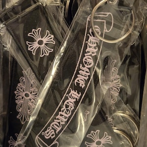 May include: A collection of black fabric keychains individually wrapped in clear plastic. Each rectangular keychain features the white embroidered text CHROME HEARTS and a white cross emblem. A silver metal ring is attached to each item.