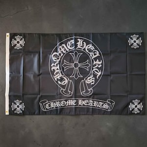 May include: A rectangular black flag featuring a large white circular emblem with a central cross and the text CHROME HEARTS. The design includes a smaller white cross in each of the four corners and a banner at the bottom with the text CHROME HEARTS.