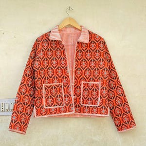 May include: A reversible jacket with a vibrant red and black leaf pattern. The jacket has two front pockets and a striped collar and lining. The jacket is hanging on a wooden hanger.