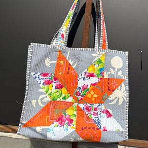 Peut inclure: Sac fourre-tout fait main avec un motif d'étoile. L'étoile est composée de tissu orange, blanc et floral. Le sac a des côtés gris avec un motif floral et des anses rayées.