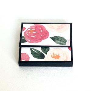 Op de afbeelding: Een kleine, zwarte doos met een bloemenontwerp. De bovenkant en binnenkant van de doos hebben een aquarelpatroon van roze en perzikrozen met groene bladeren op een witte achtergrond. De doos is open.