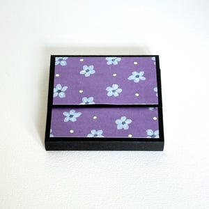 May include: A small, square black box with a hinged lid. The lid and top surface feature a purple floral pattern with light blue flowers and white dots. The box is likely for small items.