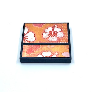 May include: A black box with a hinged lid. The box is decorated with a floral pattern of white hibiscus flowers on a yellow and orange background.