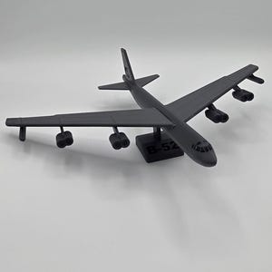 May include: A dark gray model airplane, a B-52 bomber, is displayed against a white background. The model has detailed wings, engines, and a tail. The base of the model has the text "B-52".