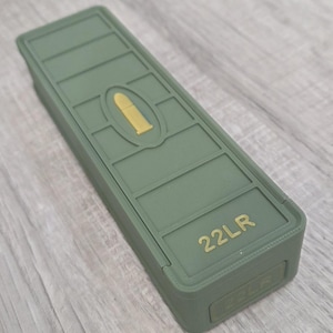 May include: An olive green rectangular container with a segmented top. A gold-colored bullet shape is centered on the lid. The text "22LR" is printed in gold on the front of the container.