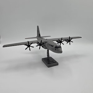 May include: A gray C-130 Hercules model airplane with four propellers and the text "U.S. AIR FORCE" on the side. The model is mounted on a gray base with the text "C-130 Hercules".