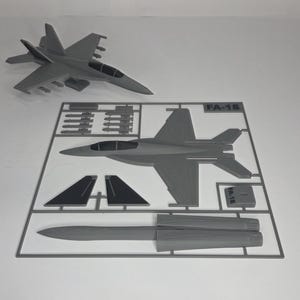 May include: A gray plastic model kit of an FA-18 fighter jet. The kit includes the assembled jet and unassembled parts on a sprue, including wings, missiles, and other components. The assembled jet is approximately 10 inches long.