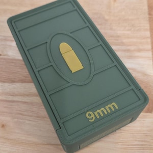 May include: A green rectangular container with a gold bullet graphic in the center. The text "9mm" is printed in gold on the front. The container is made of a matte material.