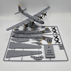 May include: A gray B-29 Superfortress model airplane, assembled and unassembled, with black propellers and yellow accents. The model includes various parts on a gray plastic frame, with the text "B-29 Super Fortress" on a small sign.