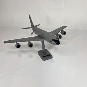 May include: A gray model airplane with four engines, wings, and a tail fin. The model is mounted on a gray stand. The text "USAF" is visible on the side of the plane. The model is a KC-135R.