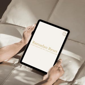 May include: A black tablet displaying the words "Ramadan Reset" in gold lettering, held by hands. The tablet also features a crescent moon and star icon. The background is a neutral, off-white color.