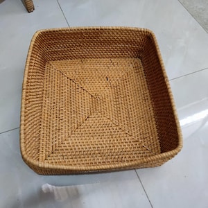 May include: A square, woven rattan basket in a natural, light brown hue. The basket has a slightly raised edge and a textured surface. The base of the basket features a woven pattern that converges towards the centre. The basket is empty.
