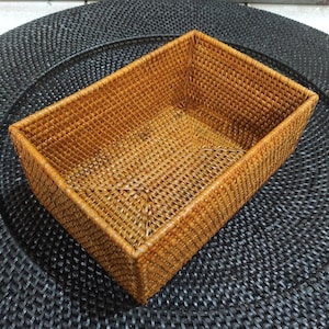 Puede incluir: Una cesta rectangular tejida en un tono marrón cálido. La cesta está hecha de fibras naturales tejidas apretadamente, con forma rectangular y lados ligeramente ensanchados. Se encuentra sobre un salvamanteles tejido negro, creando un contraste de texturas y colores.