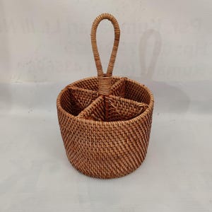 以下が含まれることがあります： 4つのコンパートメントとハンドルが付いた、丸い織りの籐製カトラリーホルダー。 天然の茶色のバスケットは、カトラリーやその他のアイテムを収納するように設計されています。 ハンドルは約18cmです。