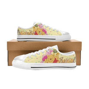 May include: Low-top canvas sneakers featuring a light yellow background with a colorful Winnie the Pooh character print, white laces, white rubber toe caps, and white soles with a thin black stripe. The text Winnie the Pooh is visible.