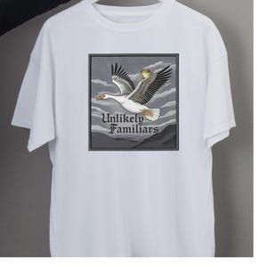Puede incluir: Camiseta blanca con un gráfico detallado de un ganso en vuelo, llevando una rana. El gráfico está enmarcado con un borde gris e incluye el texto "Unlikely Familiars". El diseño está sobre un fondo de nubes y montañas estilizadas.