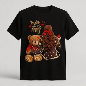 Puede incluir: Camiseta negra con un gráfico de una niña con un lazo rojo, sosteniendo una taza y un oso de peluche. El texto "Make your own magic" está encima de la niña y el oso, con acentos de corazón rojo.