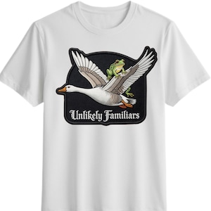 Puede incluir: Camiseta blanca con un gráfico de un ganso que lleva una rana verde, con el texto "Unlikely Familiars" en un emblema negro. El ganso tiene plumas blancas y un pico marrón. La rana es verde con ojos negros.