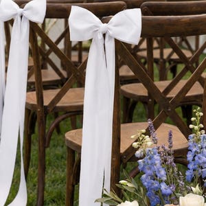 May include: Wooden chairs with white bows and ribbons are arranged for an outdoor event. The chairs have a dark wood finish and are decorated with large white bows. Floral arrangements with roses and other flowers are at the base of the chairs.