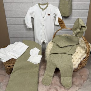 Könnte beinhalten: Eine Kollektion von Babykleidung und Accessoires in sanften, neutralen Farbtönen. Das Set umfasst einen weißen Strampler mit braunen Details, einen Lätzchen, einen Hut, eine Hose, eine Decke und Fäustlinge. Die Artikel sind auf einem rosa Teppich und in einem Weidenkorb angeordnet.