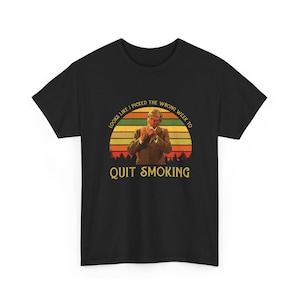 May include: Black t-shirt featuring a retro sunset design with a person holding a cigarette. The text reads "LOOKS LIKE I PICKED THE WRONG WEEK TO QUIT SMOKING" in a vintage font.
