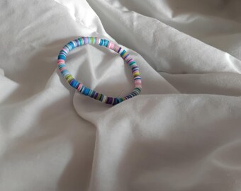 May include: A colorful bracelet made of small, flat, round beads. The beads are in shades of blue, pink, green, purple, and white. The bracelet is resting on a white, textured surface.