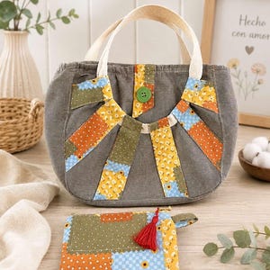 Op de afbeelding: Een grijze stoffen handtas met twee crèmekleurige hengsels en een centrale groene knoopsluiting. De tas heeft een zonnestraalpatroon van patchwork-stofstroken in gele, oranje en groene bloemenprints. Inclusief een bijpassend rechthoekig tasje met rits.
