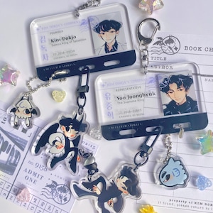 May include: Clear acrylic keychains featuring anime-style character art. The keychains have a rectangular ID card design with character portraits and text. Additional charms include cartoon figures and star-shaped embellishments. The keychains are attached to metal clasps.