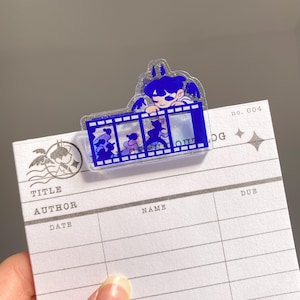 Puede incluir: Un marcapáginas de acrílico azul con un diseño de personaje de dibujos animados y tira de película. El marcapáginas está sujeto a una tarjeta de biblioteca con las palabras "TITLE", "AUTHOR", "NAME" y "DATE" impresas. La tarjeta también tiene el número "no. 004" impreso.