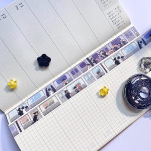 May include: Open notebook with grid paper, featuring decorative washi tape with anime-style illustrations of people in a train. Includes a black flower-shaped magnet, a yellow star-shaped magnet, and a decorative watch.