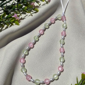 May include: A beaded phone charm featuring alternating pink and light green beads, interspersed with smaller white beads. The charm has a white cord loop for attachment. The background is a neutral fabric with floral accents.