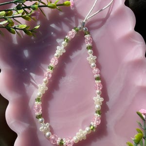Puede incluir: Un charm de teléfono con cuentas con un delicado diseño floral. El charm está compuesto por cuentas rosas, blancas y verdes, incluyendo pequeñas cuentas en forma de flor y perlas de imitación. Se adjunta un cordón blanco para colgar.