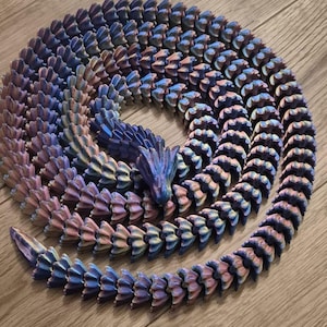 Puede incluir: Un dragón articulado impreso en 3D en tonos de azul, morado y rosa, enrollado sobre una superficie de madera. La cabeza del dragón es visible, y el cuerpo está hecho de segmentos entrelazados, creando una apariencia flexible y texturizada.