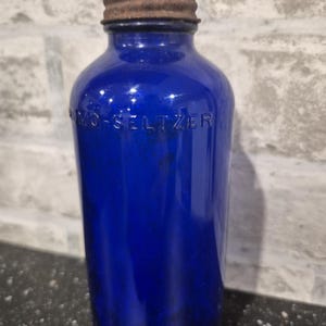 May include: A vintage cobalt blue glass bottle with a rusty metal cap. The bottle is embossed with the text "Bromo-Seltzer". The cylindrical bottle is a deep blue colour.