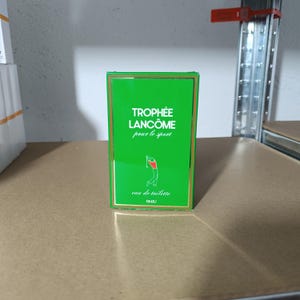 Può includere: Una scatola verde di eau de toilette Lancôme Trophée. La scatola ha bordi dorati e le parole "Trophée Lancôme pour le sport" in bianco. Un'illustrazione rossa e bianca di un golfista è sul davanti. Sono visibili anche le parole "eau de toilette" e "PARIS".