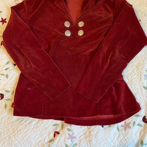 May include: A long-sleeved, burgundy velvet top with a collared neckline and four silver-colored buttons. The top has a fitted waist and is made of a soft, plush material. The garment is displayed on a floral patterned quilt.