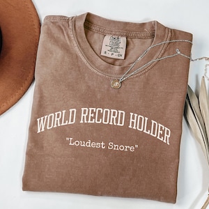May include: A brown t-shirt with the text "WORLD RECORD HOLDER" and "Loudest Snore" printed in white. A silver necklace with a pendant is draped across the shirt. A brown hat and dried palm leaf are in the background.