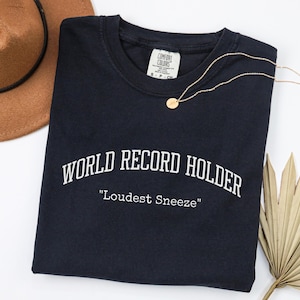 May include: A navy blue t-shirt with the text "WORLD RECORD HOLDER" and "Loudest Sneeze" in white. A gold necklace and a brown hat are also in the image. A dried palm leaf is in the bottom right corner.