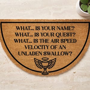 May include: A semi-circular tan coir doormat featuring black text that reads: WHAT... IS YOUR NAME? WHAT... IS YOUR QUEST? WHAT... IS THE AIR SPEED VELOCITY OF AN UNLADEN SWALLOW? Below the text is a black graphic of a winged chalice with a halo.