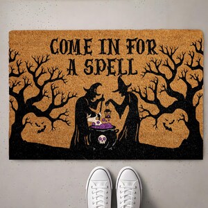 May include: A rectangular tan coir doormat featuring a black graphic of two witches stirring a cauldron with a skull design. The text reads COME IN FOR A SPELL in black lettering, surrounded by silhouettes of bare trees and small flying bats.