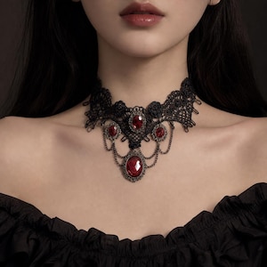 May include: A black lace choker necklace featuring intricate floral patterns and five oval-shaped red gemstones set in dark metal frames. The design includes delicate metal chains connecting the stones, creating a layered, dangling effect.